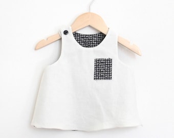 May include: A white sleeveless top with a black and white patterned pocket. The top is hanging on a wooden hanger.