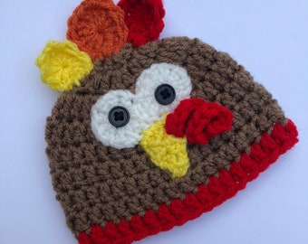 Puede incluir: Un gorro de punto marrón con un diseño de pavo. El gorro tiene un pico rojo y amarillo, ojos blancos con botones negros y una barba roja, naranja y amarilla.