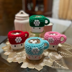 May include: Four crocheted mug cozies in red, green, pink, and blue with white snowflake embellishments. Each cozy is on a white mug with a smiling face.