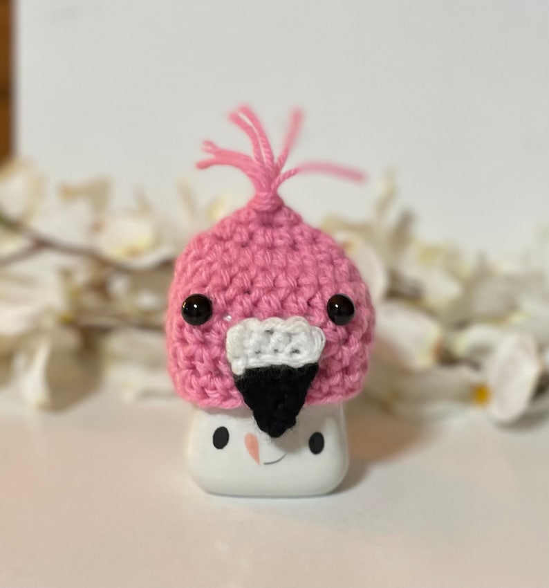 May include: A pink crocheted flamingo hat with black eyes and a black beak. The hat is sitting on a white ceramic object with a smiley face.