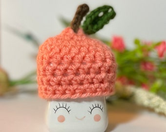 May include: A crocheted peach-colored hat with a green leaf on top, sitting on a white ceramic planter with a smiling face.