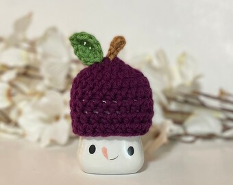 May include: A purple crocheted hat shaped like a plum with a green leaf on top. The hat is sitting on a white ceramic figurine with a smiling face.