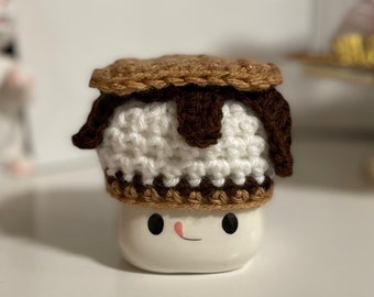 May include: A white ceramic mug with a smiling face, wearing a crocheted s'mores hat. The hat is brown, white, and dark brown, resembling a graham cracker, marshmallow, and chocolate.