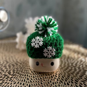 May include: A whimsical, handcrafted marshmallow figure wearing a green crocheted hat with a white and green pom-pom and white snowflake accents. The marshmallow has a smiling face. The item is on a gold textured surface.