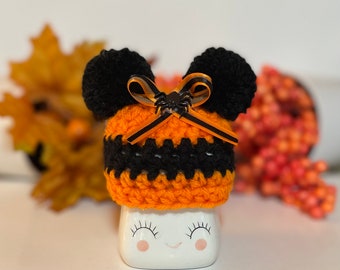 May include: A small orange and black crocheted hat with black pom-poms and a black and orange ribbon with a spider charm. The hat is sitting on a white ceramic mug with a smiling face.