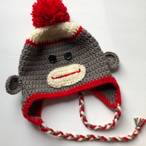 May include: Gray and red crocheted monkey hat with a white pom pom on top. The hat has a long, braided tie.