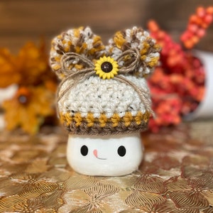 May include: A white ceramic mug with a smiling face, wearing a crocheted hat with brown, white, and yellow stripes. The hat has a brown pom-pom on top and a yellow sunflower button.