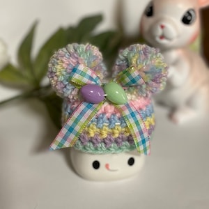 May include: A pastel rainbow crocheted hat with a plaid ribbon and pastel Easter egg beads. The hat is sitting on a white ceramic figurine with a smiling face.