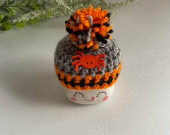 May include: A white ceramic mug with a smiling face and orange and black crocheted hat with a spider on top. The hat has a fluffy pom-pom.