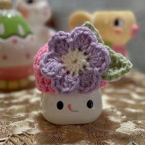 May include: A white ceramic mug with a smiling face, decorated with a crocheted flower hat in shades of pink, purple, and green.