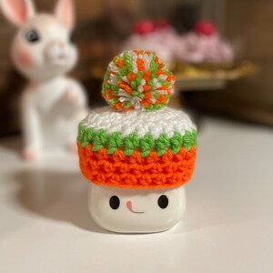 May include: A white ceramic mug with a crocheted hat. The hat is orange, green, and white with a pom-pom on top.