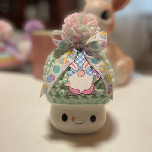 May include: A green crocheted gnome hat with a pink and white pom pom and a ribbon with Easter eggs. The hat is on top of a white ceramic mug with a smiling face.