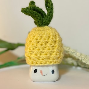 May include: A white ceramic marshmallow-shaped mug with a smiling face, wearing a yellow crocheted pineapple hat with green leaves. The mug has black eyes and a pink blush. The background is a soft white.