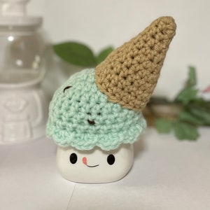May include: A crocheted ice cream cone with a light green scoop and a brown cone. The ice cream cone is sitting on a white mug with a smiling face.