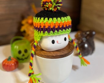 May include: A white ceramic mug with a black, green, orange, and purple crocheted hat with a pom-pom on top. The mug is sitting on a wooden lid.