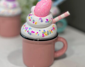 May include: A pink ceramic mug with a white whipped cream topping, pink and white sprinkles, and a pink candy egg. The mug has a black rim and a black handle. A striped straw is sticking out of the whipped cream.