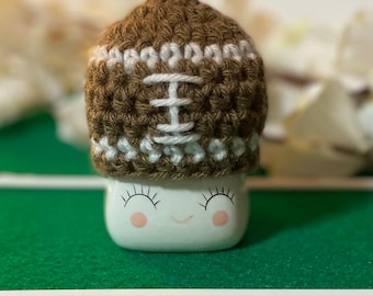 May include: A brown and white crocheted football hat sits on top of a white marshmallow with a smiling face. The marshmallow is on a green surface with a white line.