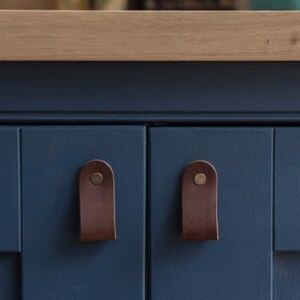 May include: A close-up shot of a navy blue cabinet with two doors. Each door features a brown leather pull secured with a brass rivet. The cabinet is topped with light wood. The pulls offer a simple, modern aesthetic.
