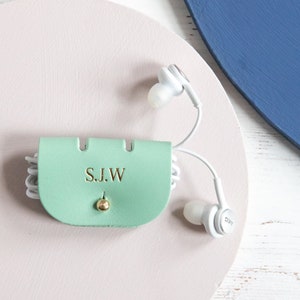 May include: A mint green leather earbud case with the initials "S.J.W" embossed in gold. The case is secured with a gold-coloured stud and holds white earbuds. The case is on a pink surface with blue and white accents.