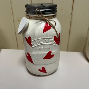 May include: A white mason jar decorated with red hearts, a silver lid, and a jute twine bow. A small white heart-shaped tag hangs from the jar. The jar has a slot for coins and is likely a money box.