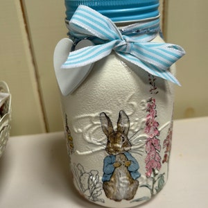 May include: A white ceramic jar with a blue lid and a blue and white striped ribbon. The jar features a printed image of Peter Rabbit from Beatrix Potter's children's book.