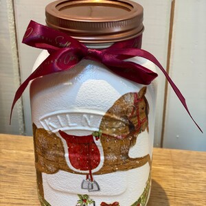 May include: A white jar with a bronze lid and a burgundy ribbon tied around the neck. The jar is decorated with a brown rocking horse image and gift boxes. A festive holiday decoration.