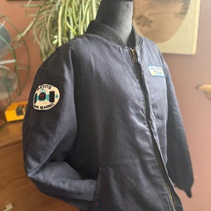 May include: Navy blue bomber jacket with a black collar and a gold zipper. The jacket features two patches: one with the text "LEVITE" and "77" and the other with the text "DRAG RUN TEAM". The jacket has two front pockets.