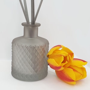 May include: A grey frosted glass diffuser bottle with a diamond pattern and grey reeds. A single yellow tulip with red tips is next to the bottle.