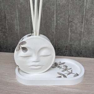 May include: White ceramic diffuser with a moon face design and a tray with a eucalyptus leaf pattern. The diffuser has a round base and a cylindrical neck with sticks inserted into it.