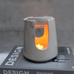 Pode incluir: Queimador de &oacute;leo de cer&acirc;mica branca com um design de m&aacute;rmore. O queimador tem uma pequena tigela no topo e um recorte no meio para uma vela de ch&aacute;. O queimador est&aacute; aceso e a vela brilha.