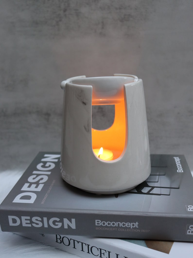 Pode incluir: Queimador de &oacute;leo de cer&acirc;mica branca com um design de m&aacute;rmore. O queimador tem uma pequena tigela no topo e um recorte no meio para uma vela de ch&aacute;. O queimador est&aacute; aceso e a vela brilha.