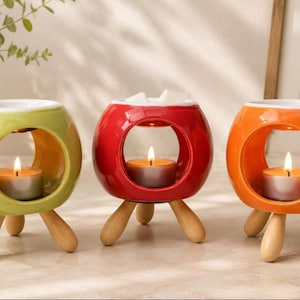 May include: Three brightly coloured ceramic wax melt burners in green, red, and orange. Each has a circular opening and wooden tripod legs. A lit tea light candle sits inside each, with wax melts in the red one.