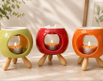 May include: Three brightly coloured ceramic wax melt burners in green, red, and orange. Each has a circular opening and wooden tripod legs. A lit tea light candle sits inside each, with wax melts in the red one.