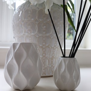 May include: Three white ceramic vases with a textured design. The vase on the left has a wavy pattern, the vase in the centre has a more intricate pattern, and the vase on the right has a wavy pattern. The vase in the centre is holding a bouquet of white lilies.