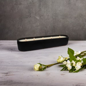May include: A black rectangular candle holder with five lit candles. The candle holder is on a white wooden surface with white flowers.