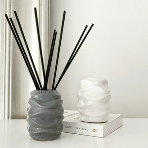 May include: Two ceramic diffuser bottles, one grey and one white, with black diffuser sticks in the grey bottle. The bottles have a wavy, textured design.