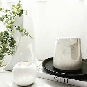Pode incluir: Aquecedor de velas de cer&acirc;mica branca com design de m&aacute;rmore, um porta-velas de cer&acirc;mica branca e um vaso branco com folhas verdes. O aquecedor de velas est&aacute; sobre uma bandeja preta e um livro com a palavra "WHITE" impressa na capa.