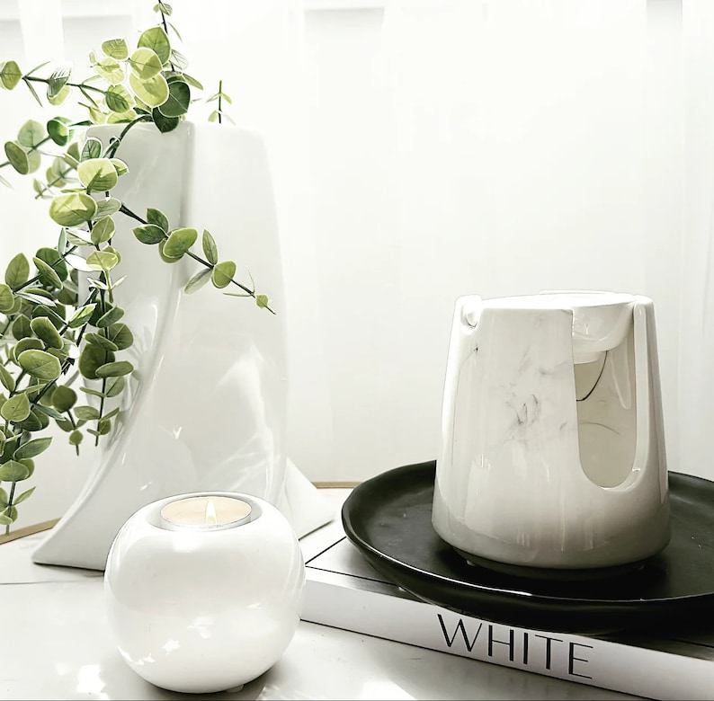 Pode incluir: Aquecedor de velas de cer&acirc;mica branca com design de m&aacute;rmore, um porta-velas de cer&acirc;mica branca e um vaso branco com folhas verdes. O aquecedor de velas est&aacute; sobre uma bandeja preta e um livro com a palavra "WHITE" impressa na capa.