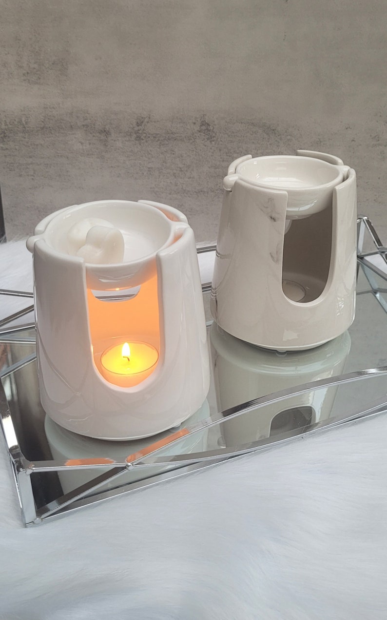 Pode incluir: Dois aquecedores de cera de cer&acirc;mica branca com uma vela de ch&aacute; em um dos aquecedores. Os aquecedores est&atilde;o em uma bandeja de espelho prateada.