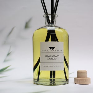 May include: A clear glass bottle diffuser with black reeds and a label that reads "Catsy Candles Lemongrass & Ginger Hand Made Fragrance Oil Reed Diffuser". The bottle is filled with a yellow liquid and has a wooden cap.