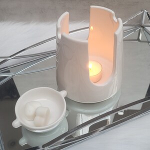 Pode incluir: Aquecedor de cera branco de cer&acirc;mica com uma pequena vela branca dentro. O aquecedor tem um design &uacute;nico com um espa&ccedil;o aberto no centro. Uma pequena tigela branca com duas ceras em forma de cora&ccedil;&atilde;o fica ao lado do aquecedor.