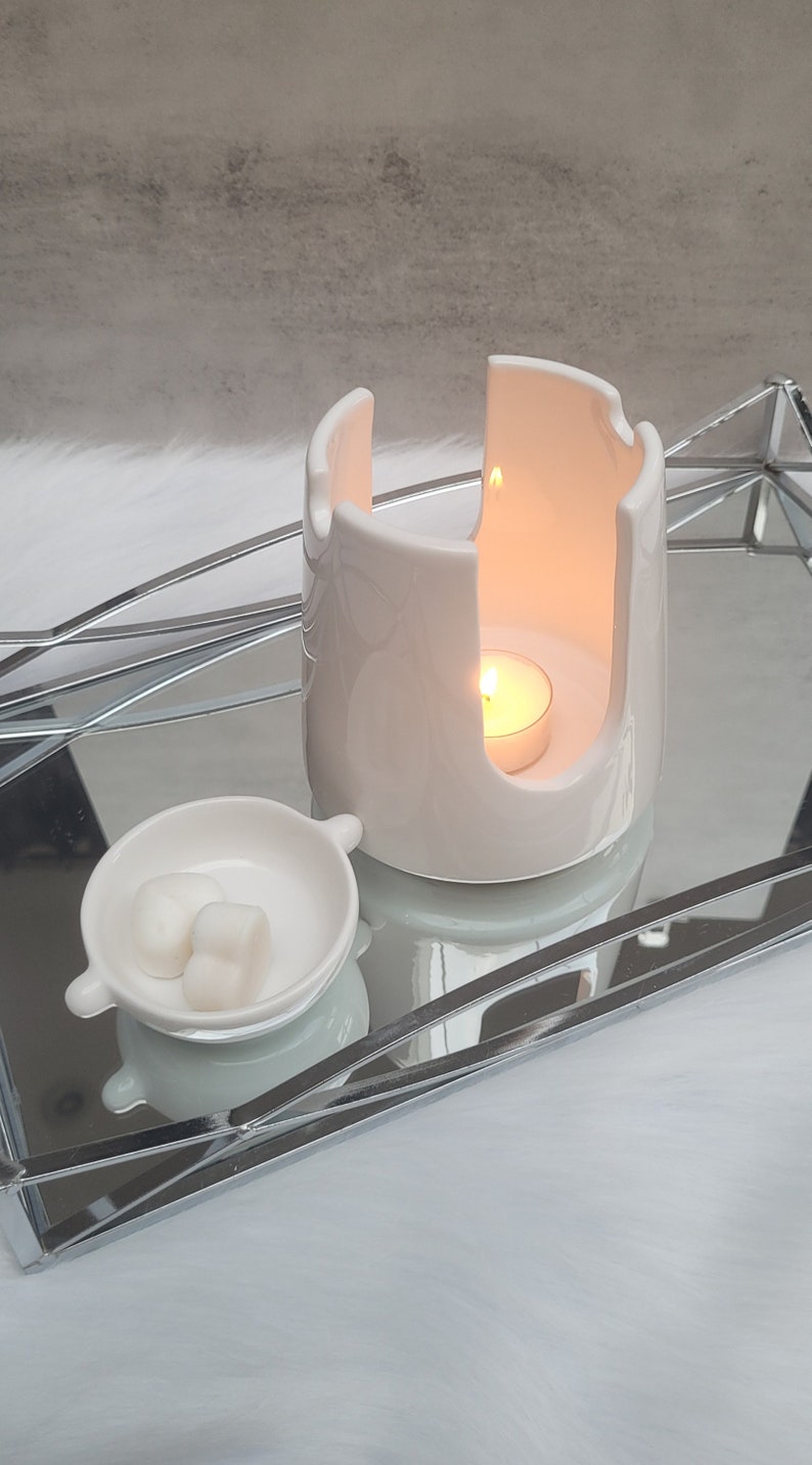 Pode incluir: Aquecedor de cera branco de cer&acirc;mica com uma pequena vela branca dentro. O aquecedor tem um design &uacute;nico com um espa&ccedil;o aberto no centro. Uma pequena tigela branca com duas ceras em forma de cora&ccedil;&atilde;o fica ao lado do aquecedor.