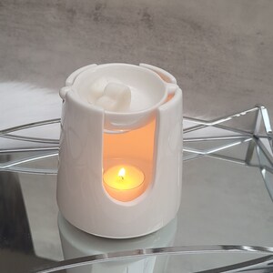 Pode incluir: Aquecedor de cera branco de cer&acirc;mica com uma pequena vela queimando dentro. O aquecedor tem uma parte superior redonda com um pequeno po&ccedil;o para derreter cera e uma se&ccedil;&atilde;o inferior com um espa&ccedil;o para a vela. O aquecedor est&aacute; em uma bandeja prateada.