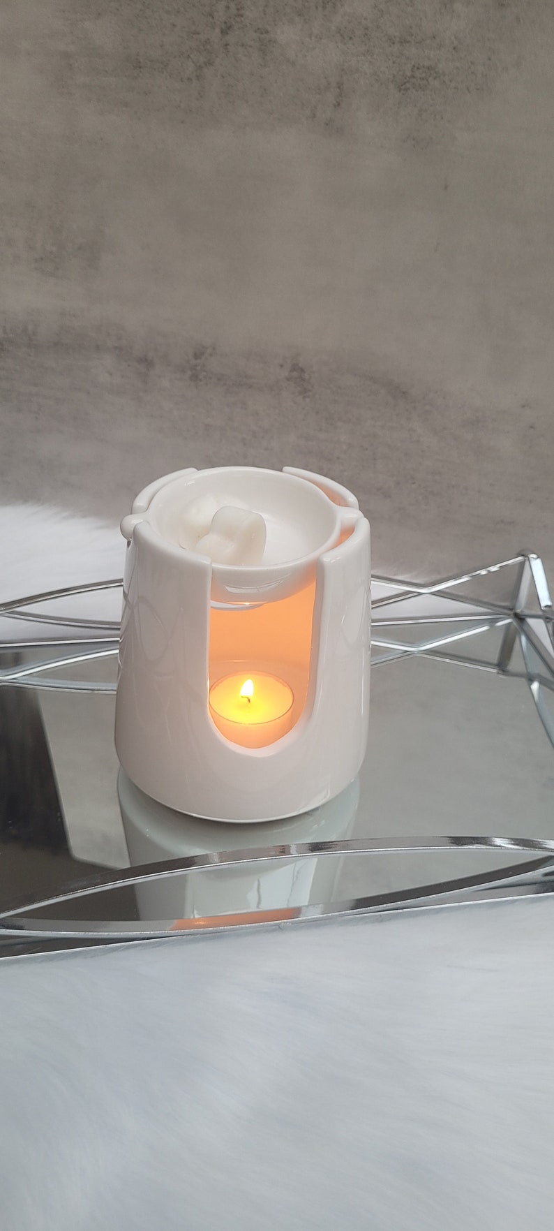 Pode incluir: Aquecedor de cera branco de cer&acirc;mica com uma pequena vela queimando dentro. O aquecedor tem uma parte superior redonda com um pequeno po&ccedil;o para derreter cera e uma se&ccedil;&atilde;o inferior com um espa&ccedil;o para a vela. O aquecedor est&aacute; em uma bandeja prateada.