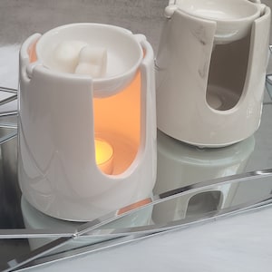 Pode incluir: Dois aquecedores de cera de cer&acirc;mica branca com um design de m&aacute;rmore. O aquecedor &agrave; esquerda tem uma vela de ch&aacute; acesa e duas ceras derretidas na tigela superior. O aquecedor &agrave; direita tem um design branco liso.
