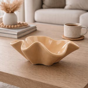 May include: A light-yellow ceramic bowl with a wavy, organic design. The bowl is placed on a light-wood table. The background features a neutral colour palette. The bowl's shape is reminiscent of a flower or seashell.