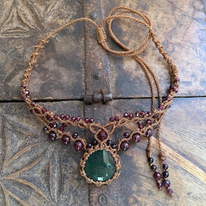May include: A brown macrame necklace with a green stone pendant and red beads. The necklace is on a wooden surface.