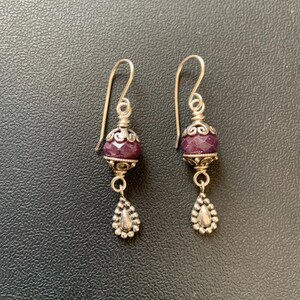 May include: A pair of silver dangle earrings. Each earring features a faceted, deep purple bead, a decorative silver cap, and a teardrop-shaped charm. The earrings have a simple hook design.