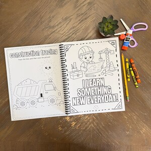 A childrens learning workbook open on a wooden table displaying a dump truck line tracing template alongside actual crayons, scissors, and a glue stick.