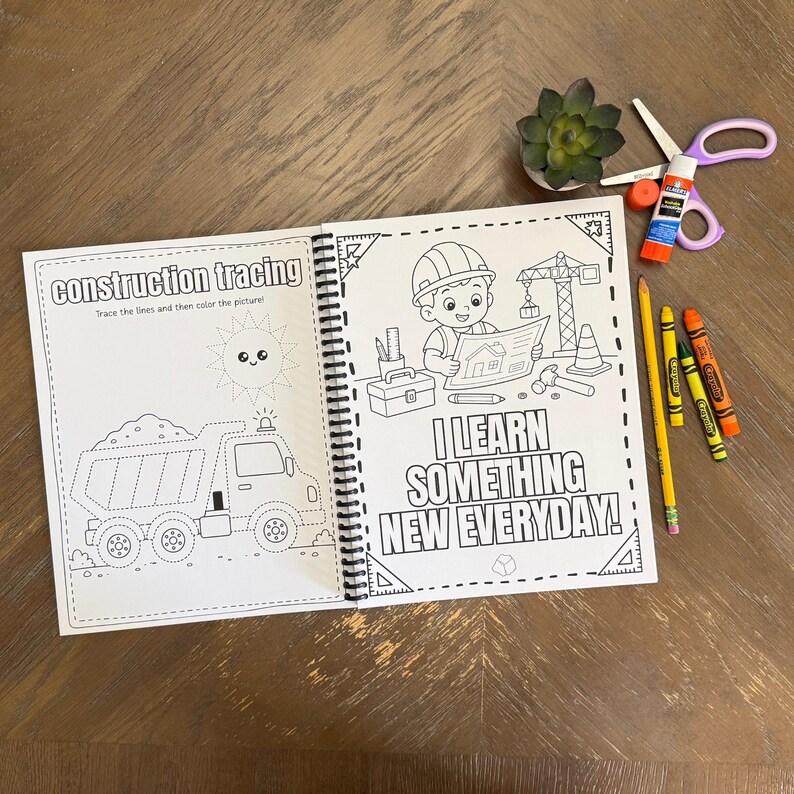 A childrens learning workbook open on a wooden table displaying a dump truck line tracing template alongside actual crayons, scissors, and a glue stick.